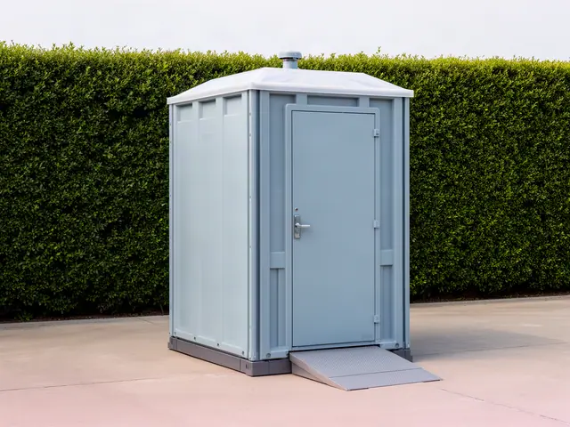 A large light grey-blue portable restroom with a low ramp at its door, standing on an empty concrete lot in front of a tall trimmed green hedge.