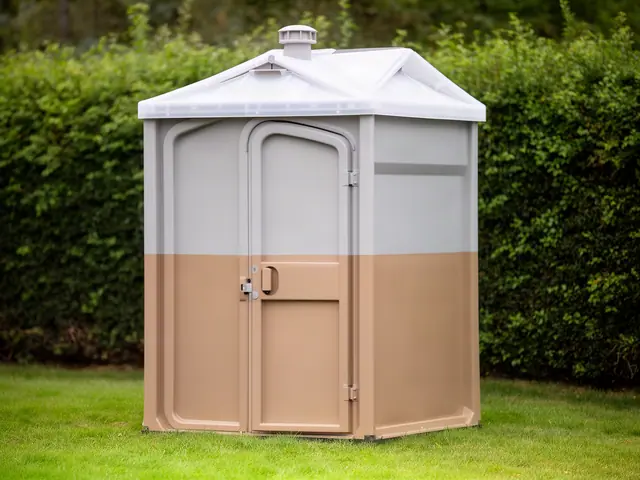 A larger portable restroom with grey upper walls, tan lower walls, a translucent peaked roof and a roof vent, standing on a lawn in front of a tall green hedge.