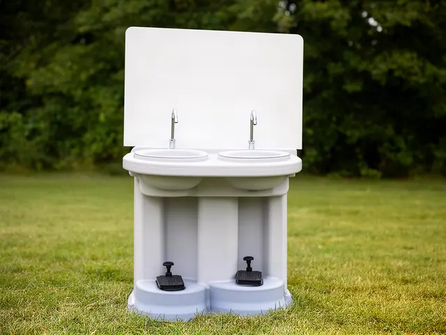 A white freestanding portable hand-washing station with two round sinks, two tall faucets and two black foot pumps at its base, standing on grass.